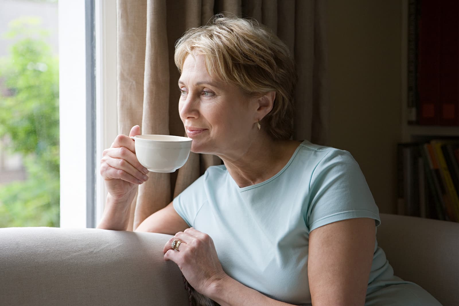 A woman in her fifties pauses by a window with a cup of tea, looking out thoughtfully.